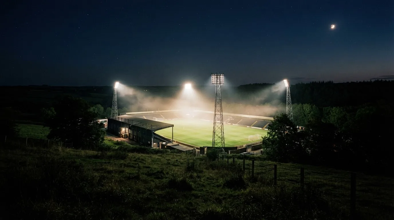 Ein Stadion unter Flutlicht bei Nacht, von einem Hügel aus der Ferne betrachtet, mit Dunst über dem Spielfeld.