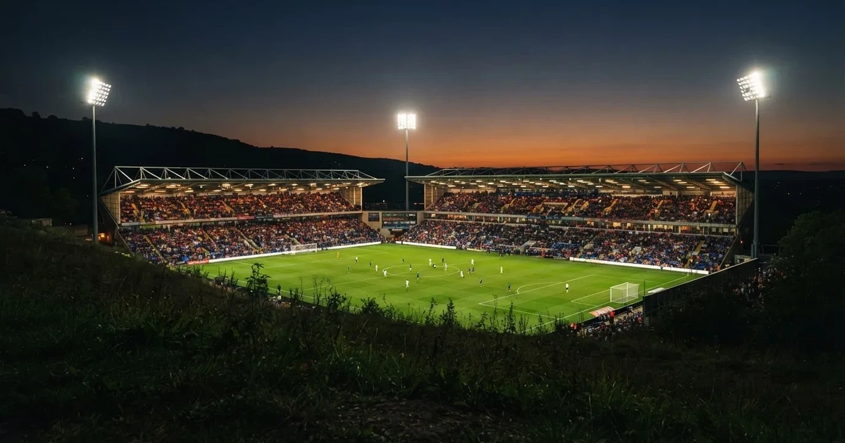Ein Fußballstadion unter Flutlicht mit hellen Rängen, von einem Hügel aus gesehen, darüber ein dunkelblauer Abendhimmel.