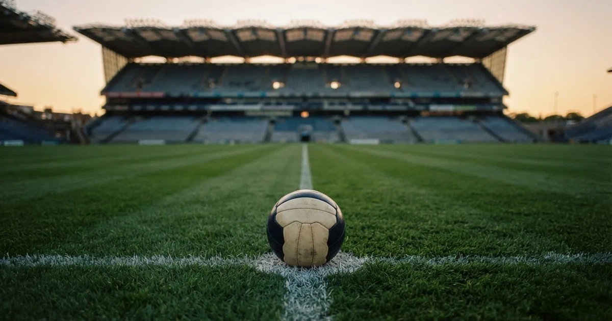 Ein Fußball liegt auf dem Anstoßpunkt eines leeren Rasenplatzes, im Hintergrund eine große Stadiontribüne im Abendlicht.