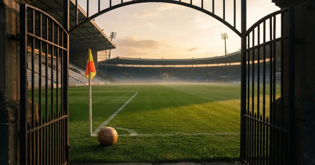 Blick durch ein Stadiontor auf einen grünen Rasen im Abendlicht, ein Ball ruht an der Eckfahne, die Tribünen sind leer.