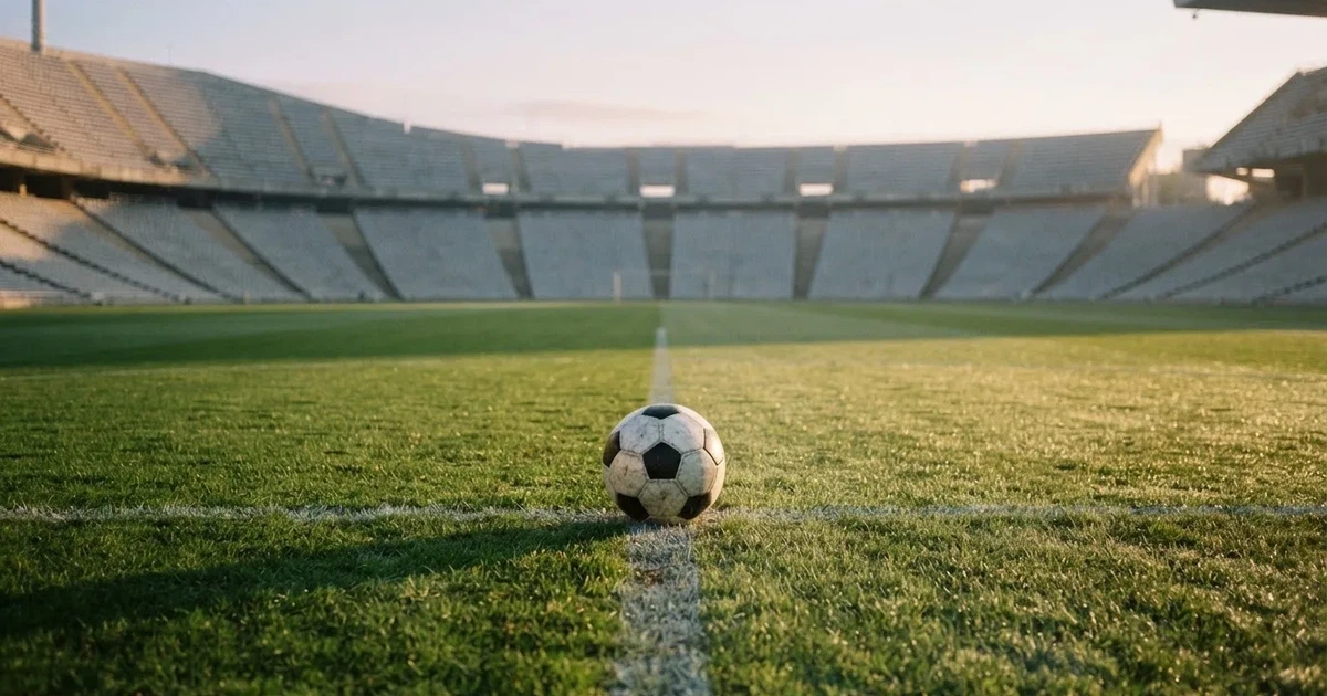 Ein Fußball liegt auf dem Mittelkreis eines Stadionrasens am frühen Morgen, im Hintergrund leere Ränge ohne erkennbare Beschriftung.