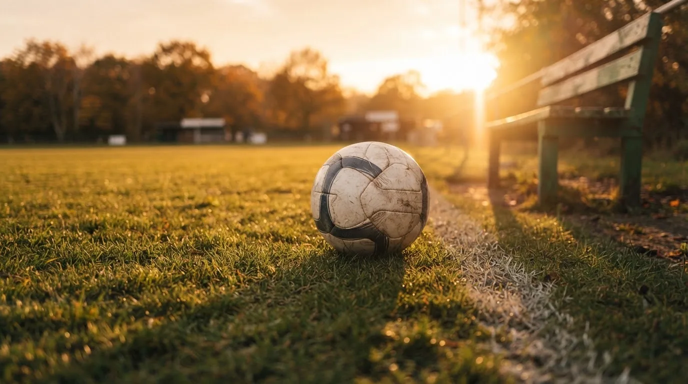 Ein Fußball auf einem Rasenplatz im Gegenlicht der tiefstehenden Sonne, am Spielfeldrand eine leere Ersatzbank.