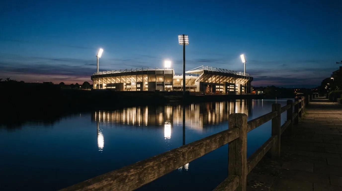 Ein Stadion unter Flutlicht aus der Ferne gesehen, im Vordergrund ein ruhiger Fluss und ein Fußweg.