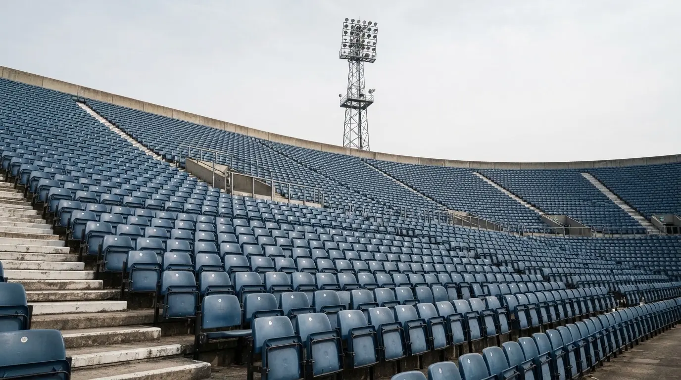 Eine leere Tribüne eines Fußballstadions bei Tageslicht, Sitzschalen in Reihen und dahinter ein Flutlichtmast.