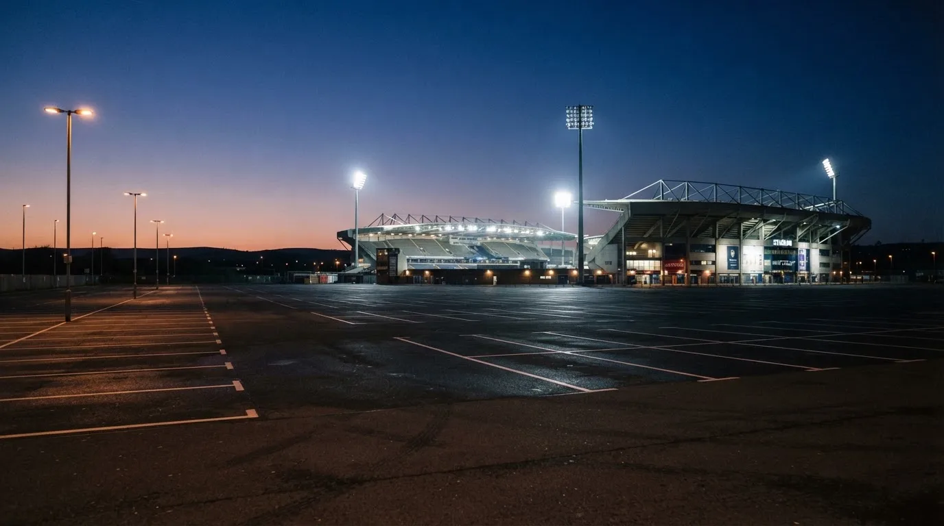 Ein Stadion unter Flutlicht aus der Ferne gesehen, davor ein leerer Parkplatz in der Dämmerung.