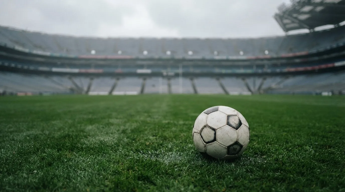 Ein Fußball auf einem Rasen, im Hintergrund ein leeres Stadion bei bewölktem Himmel.