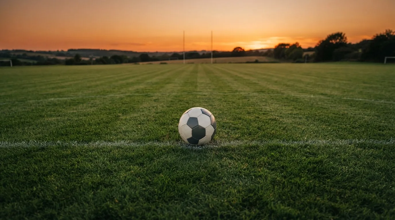 Ein Fußball liegt in der Mitte eines leeren Rasenplatzes, am Horizont färbt sich der Himmel orange.
