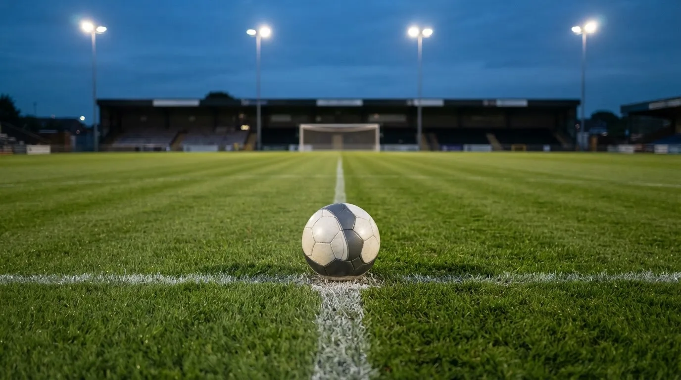 Ein Fußball liegt auf dem Anstoßpunkt eines leeren Rasenplatzes, im Hintergrund ein Stadion unter Flutlicht in der Abenddämmerung.