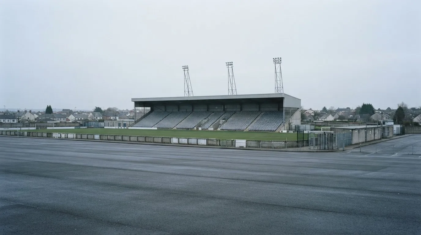 Eine leere Tribüne eines kleinen Stadions am frühen Morgen, aus der Ferne über einen menschenleeren Parkplatz fotografiert.