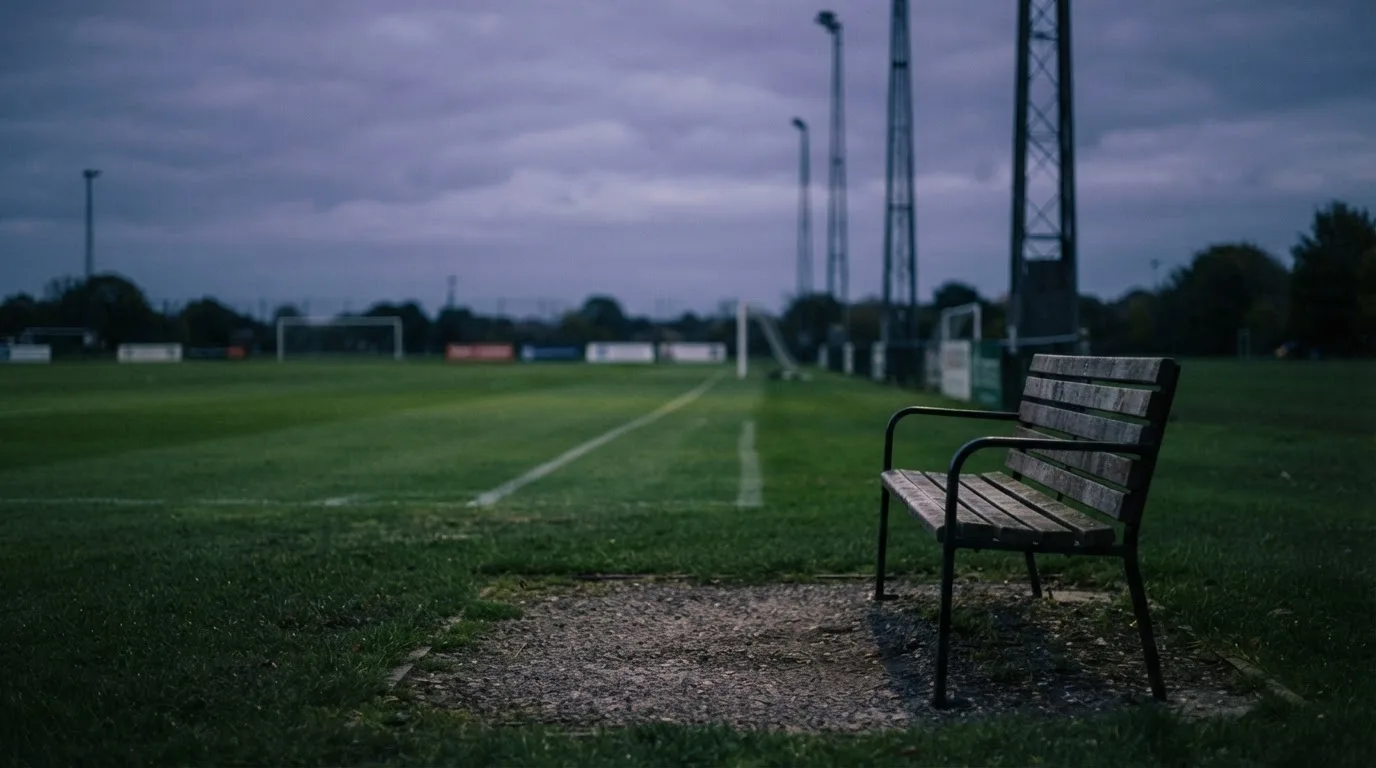 Eine leere Parkbank am Rand eines Fußballfelds in der Abenddämmerung, im Hintergrund ragen dunkle Flutlichtmasten in den Himmel.