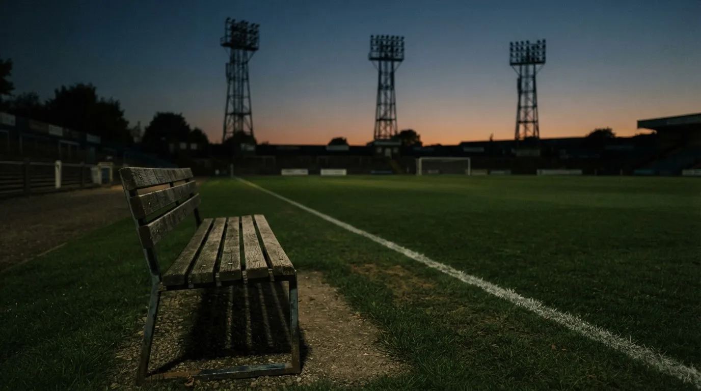 Eine leere Parkbank am Rand eines Fußballfelds in der Abenddämmerung, im Hintergrund ragen dunkle Flutlichtmasten in den Himmel.