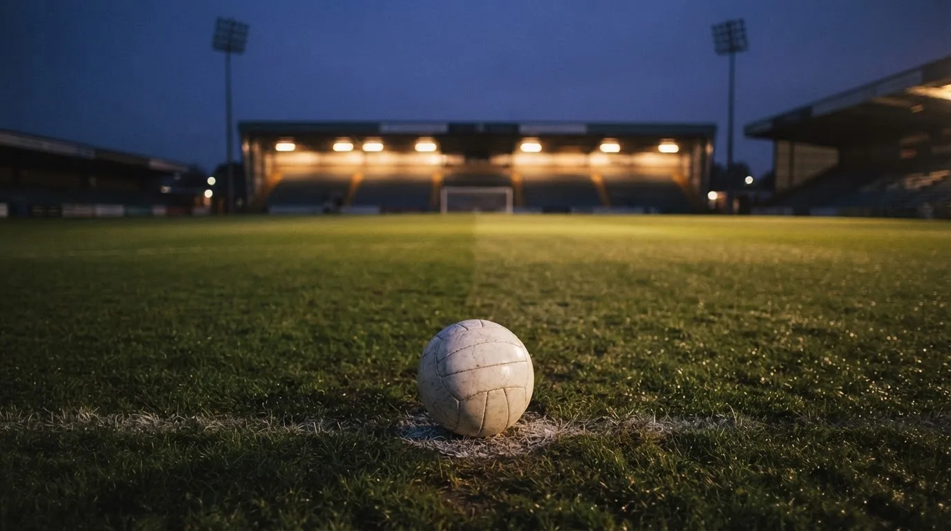 Ein Fußball liegt auf dem Anstoßpunkt eines leeren Rasenplatzes, im Hintergrund steht ein Stadion unter Flutlicht in der Dämmerung.