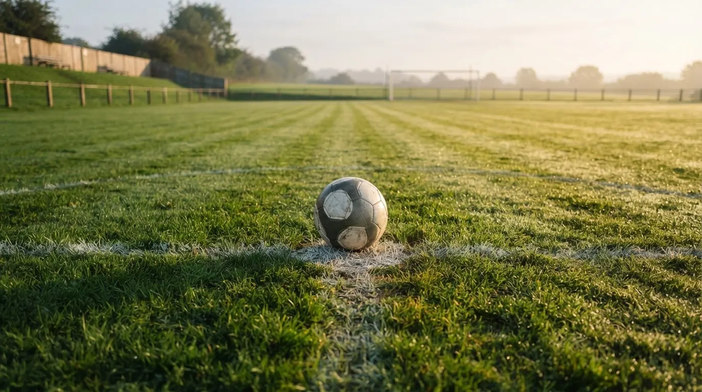 Ein Fußball liegt auf dem Anstoßkreis eines leeren Rasenplatzes im Licht der Morgensonne.