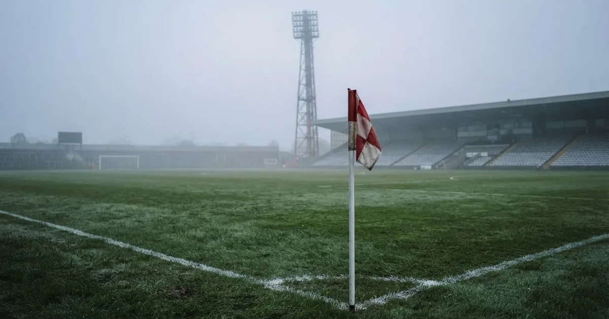 Eine Eckfahne steht im Nebel am Rand eines leeren Rasenplatzes in der Morgendämmerung, dahinter ein Flutlichtmast.