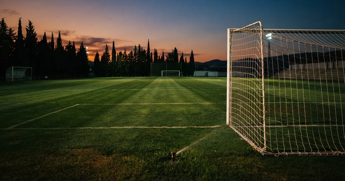 Ein Fußballtor mit Netz auf einem leeren Platz im Abendlicht, der Rasen frisch gemäht, im Hintergrund Zypressen.