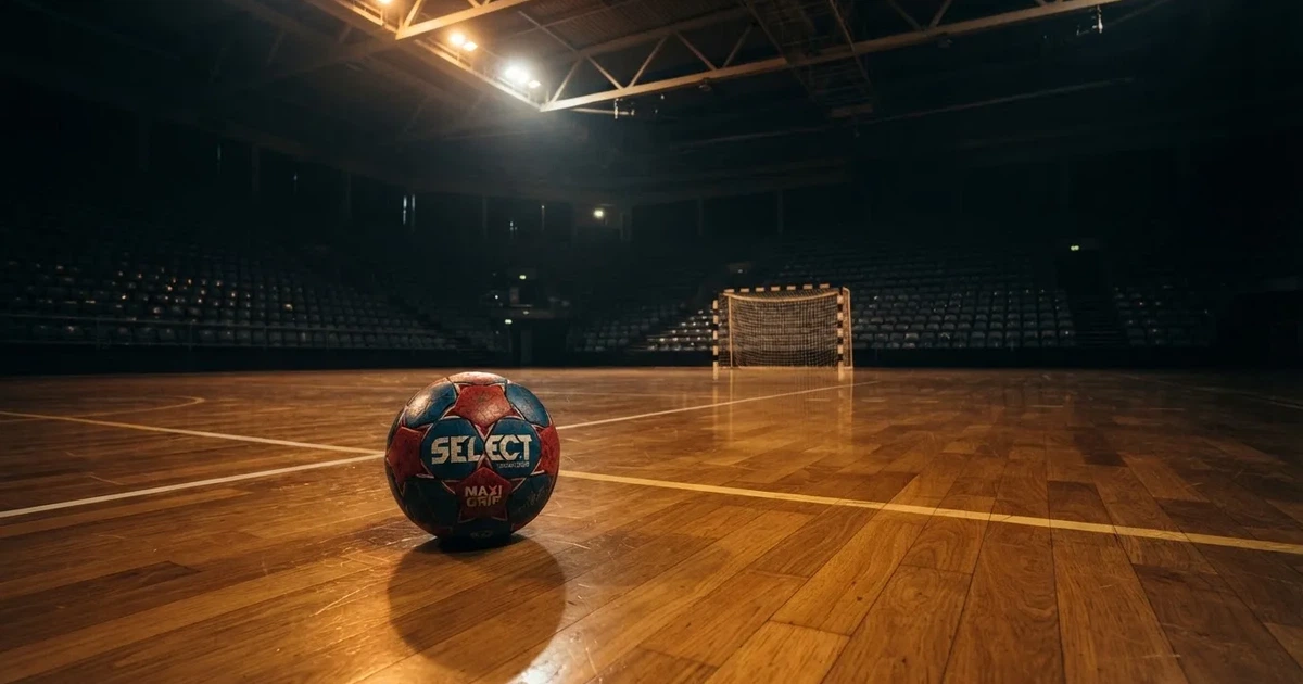 Ein Handball liegt auf dem Hallenboden vor einem Tor mit Netz, die leere Halle wird von Flutlicht beleuchtet.