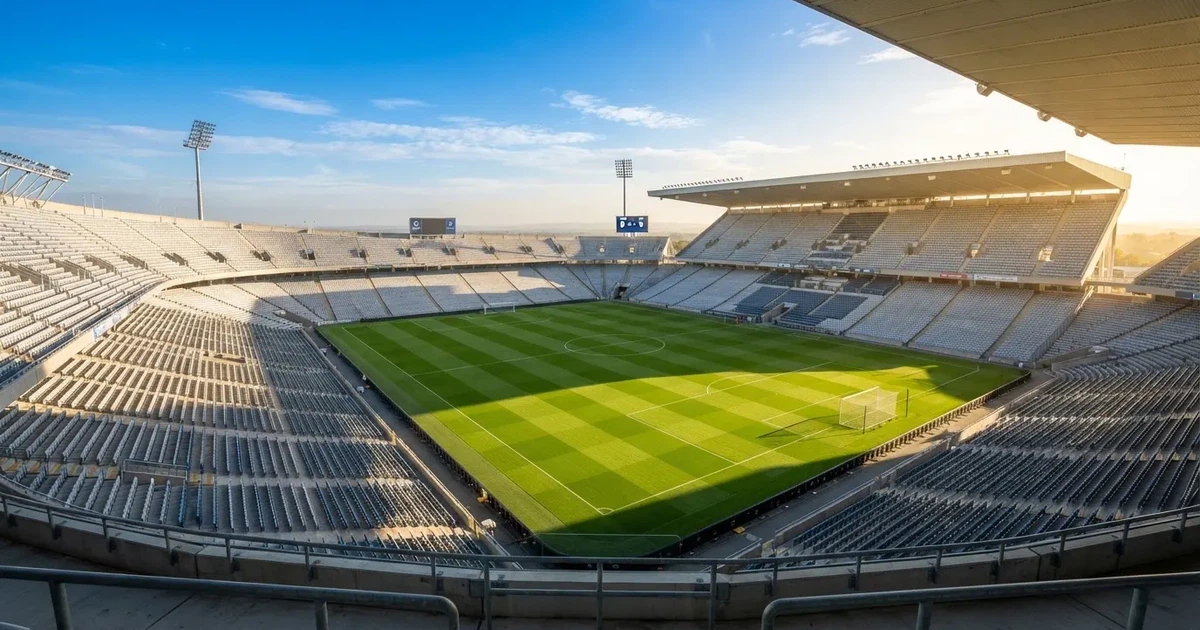 Ein Fußballstadion mit hellen Rängen unter strahlend blauem Himmel, davor ein sonnenbeschienener Rasen mit frisch gemähten Streifen.