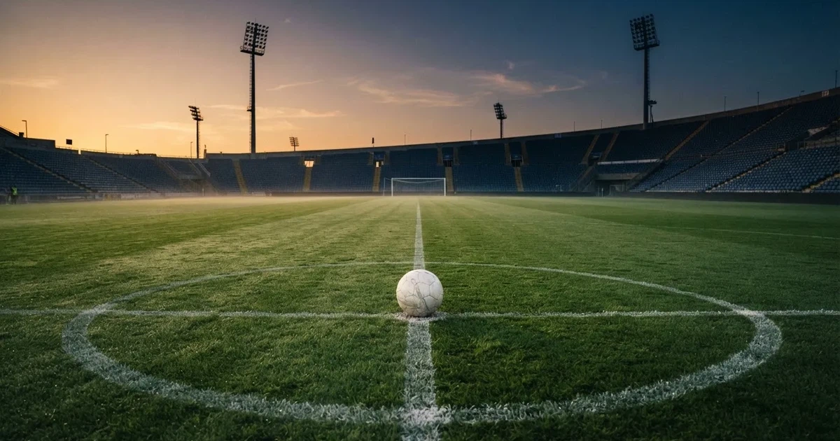Ein Fußball liegt auf dem Anstoßpunkt eines leeren Rasenplatzes, im Hintergrund eine Tribüne und Flutlichtmasten in der Abenddämmerung.