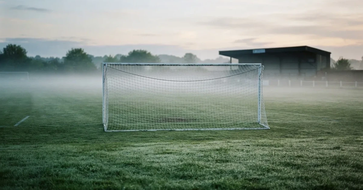Ein Fußballtor mit Netz steht im Morgennebel auf einem leeren Rasenplatz, im Hintergrund die Silhouette einer kleinen Tribüne.