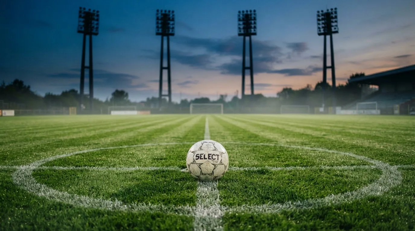 Ein Fußball liegt im Gras eines leeren Platzes, im Hintergrund stehen Flutlichtmasten in der Dämmerung.