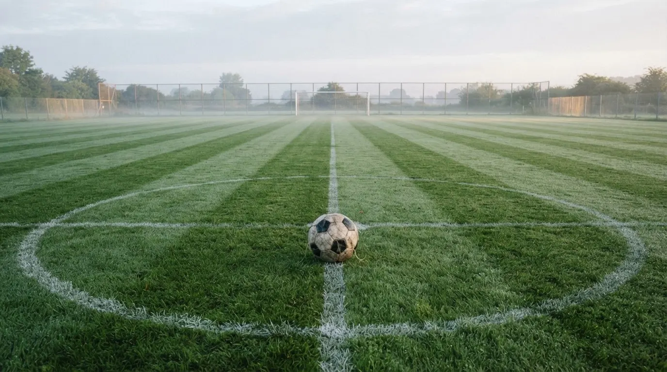 Ein Fußball liegt allein auf dem Anstoßpunkt eines leeren Rasenplatzes im Morgenlicht, am Rand ein niedriger Zaun.