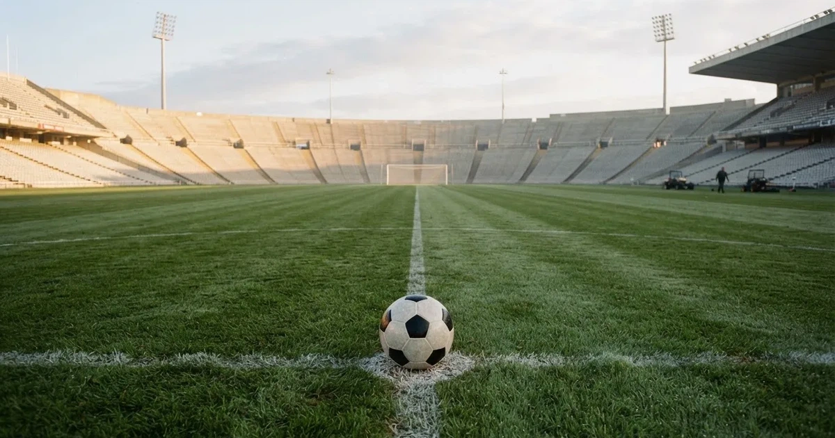 Ein Fußball liegt auf dem Anstoßpunkt eines Rasenplatzes, dahinter leere Tribünen im Licht des frühen Morgens.