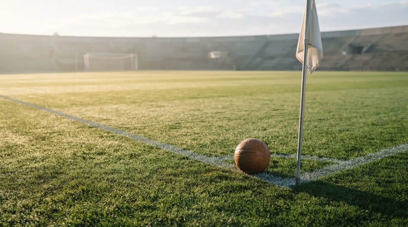 Ein Fußball liegt auf Rasen neben einer Eckfahne ohne Aufdruck auf einem leeren Platz im Morgenlicht.