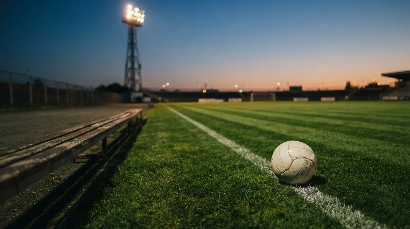 Ein Fußball liegt am Spielfeldrand neben einer leeren Ersatzbank, das Flutlicht über dem Platz leuchtet in der Dämmerung.