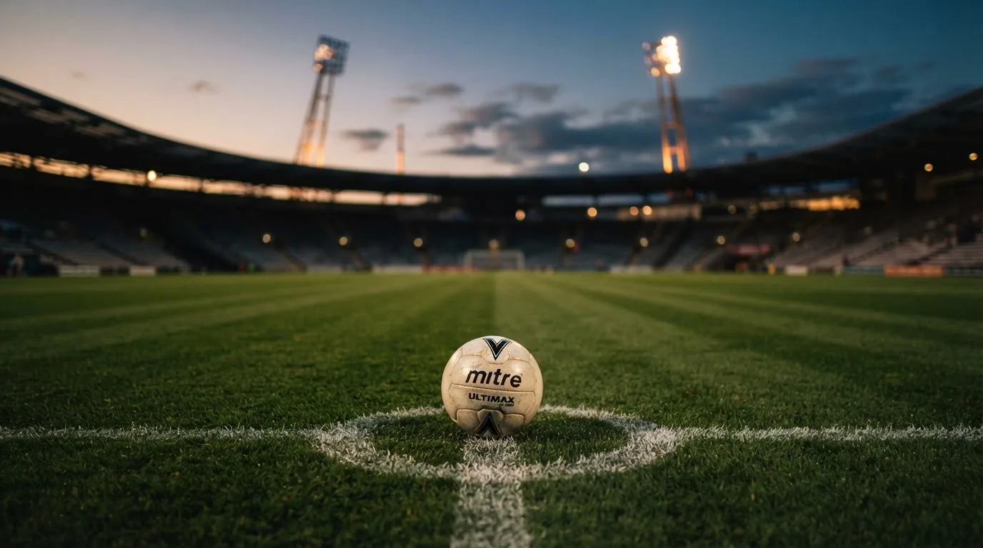 Ein Fußball liegt auf dem Anstoßpunkt eines Rasenplatzes in der Abenddämmerung, Flutlichtmasten stehen unscharf im Hintergrund.